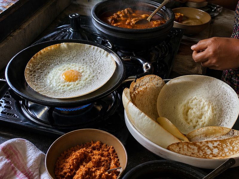 A woman preparing a traditional Sri Lankan breakfast of hoppers and curry in a home kitchen.