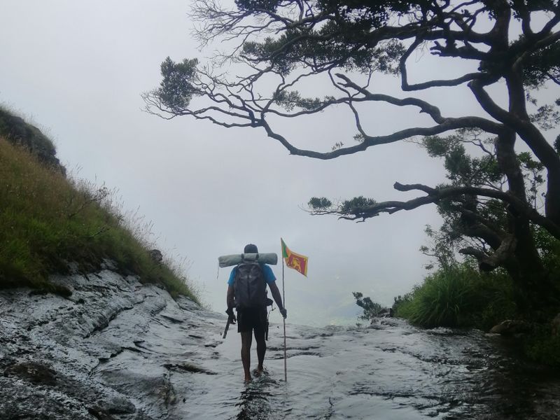 A lone hiker with a backpack crossing a river valley in a mountainous landscape.